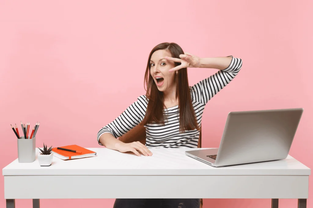 young-excited-specialist-woman-with-opened-mouth-showing-victory-sign-white-desk-with-contemporary-laptop